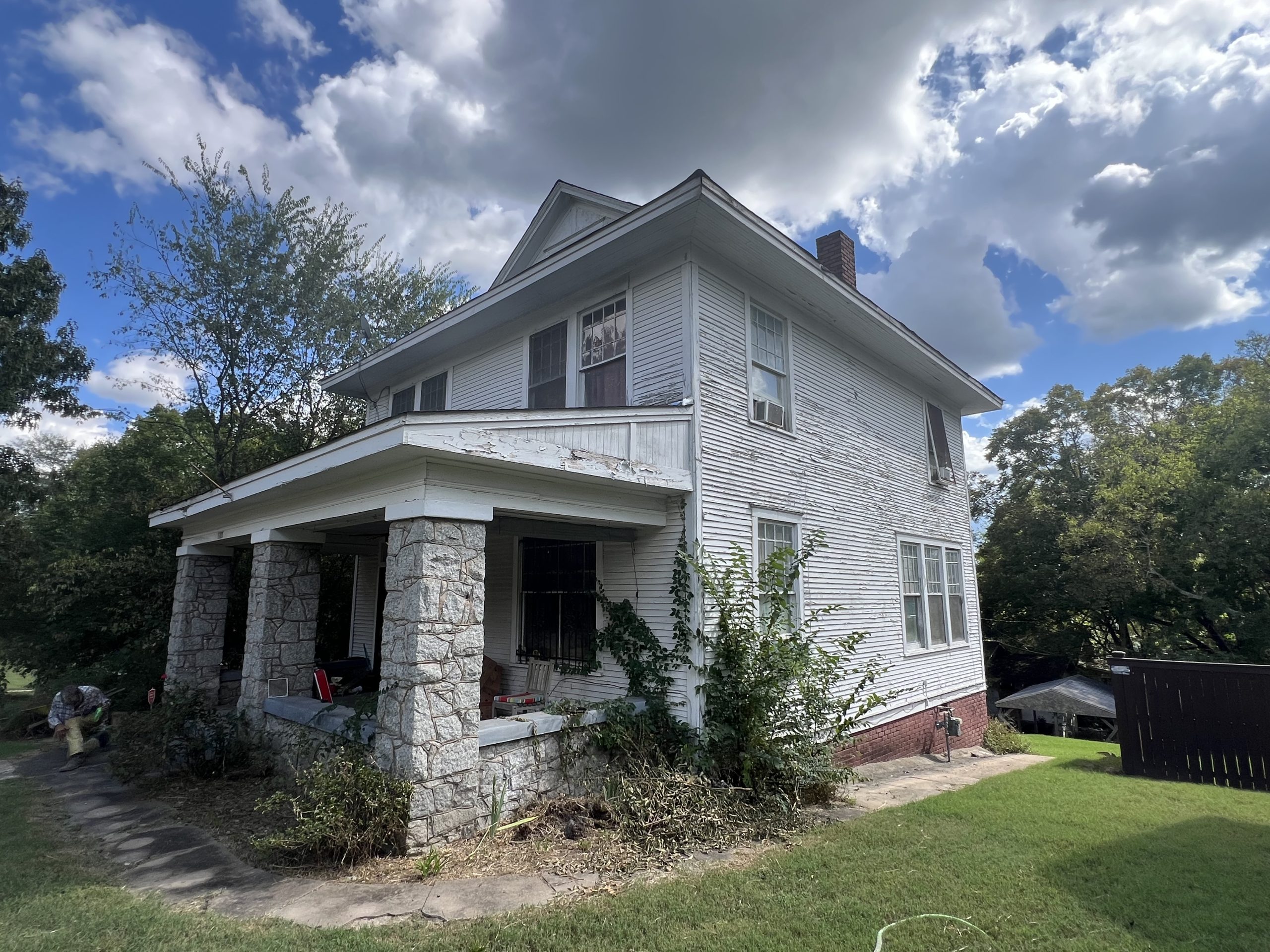 A two story white house with a covered front porch supported by stone columns, viewed from an angled side perspective. The exterior shows peeling paint and overgrown plants around the foundation, with a grassy yard, neighboring structures, and a partly cloudy sky in the background.