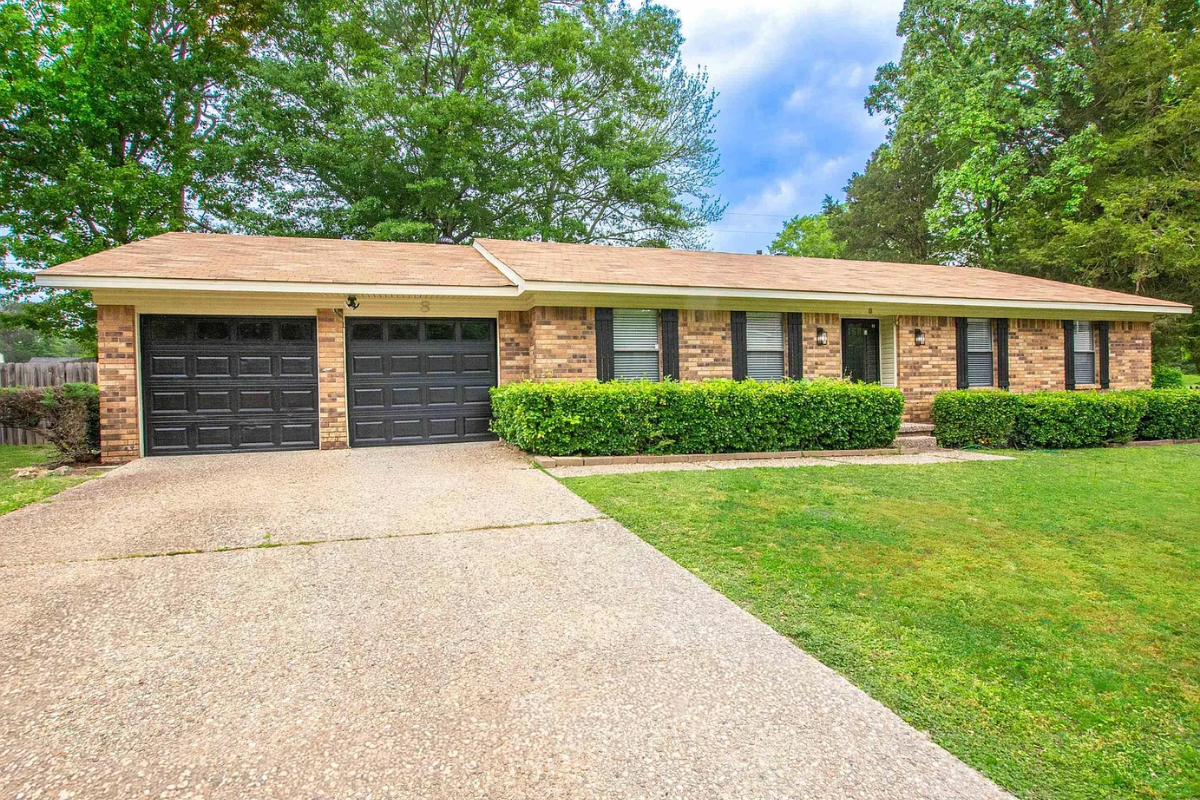 A single story brick house with a two car garage and black garage doors, viewed from the driveway. The home features neatly trimmed hedges along the front, a well maintained lawn, and large trees surrounding the property under a partly cloudy sky.